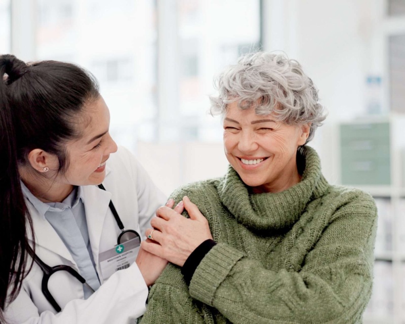 Smiling female doctor in a white coat interacting warmly with an older woman in a green sweater.