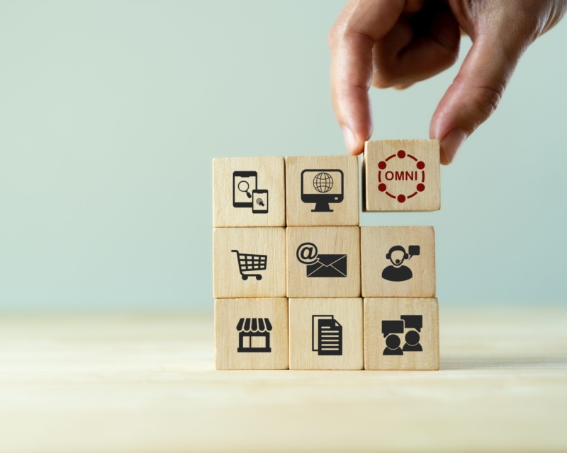 Hand placing a wooden block labeled “Omni” on top of stacked blocks with icons representing various marketing channels.
