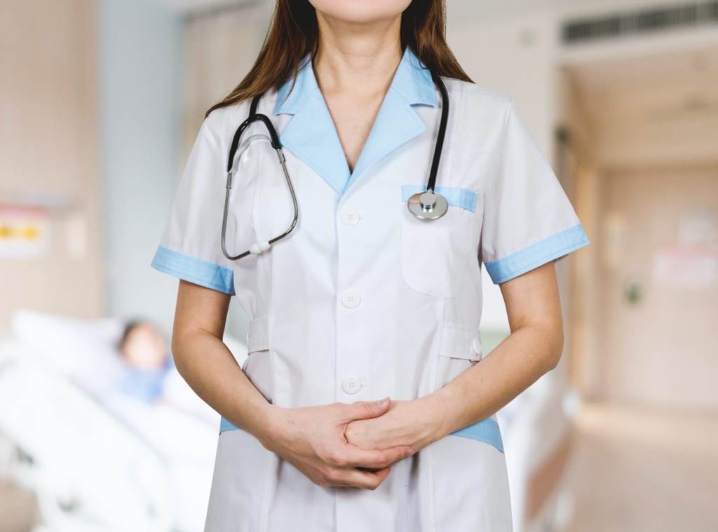 Nurse A nurse in blue scrubs stands in a hospital room, looking toward the camera. A patient is visible in a bed in the background, which is out of focus.