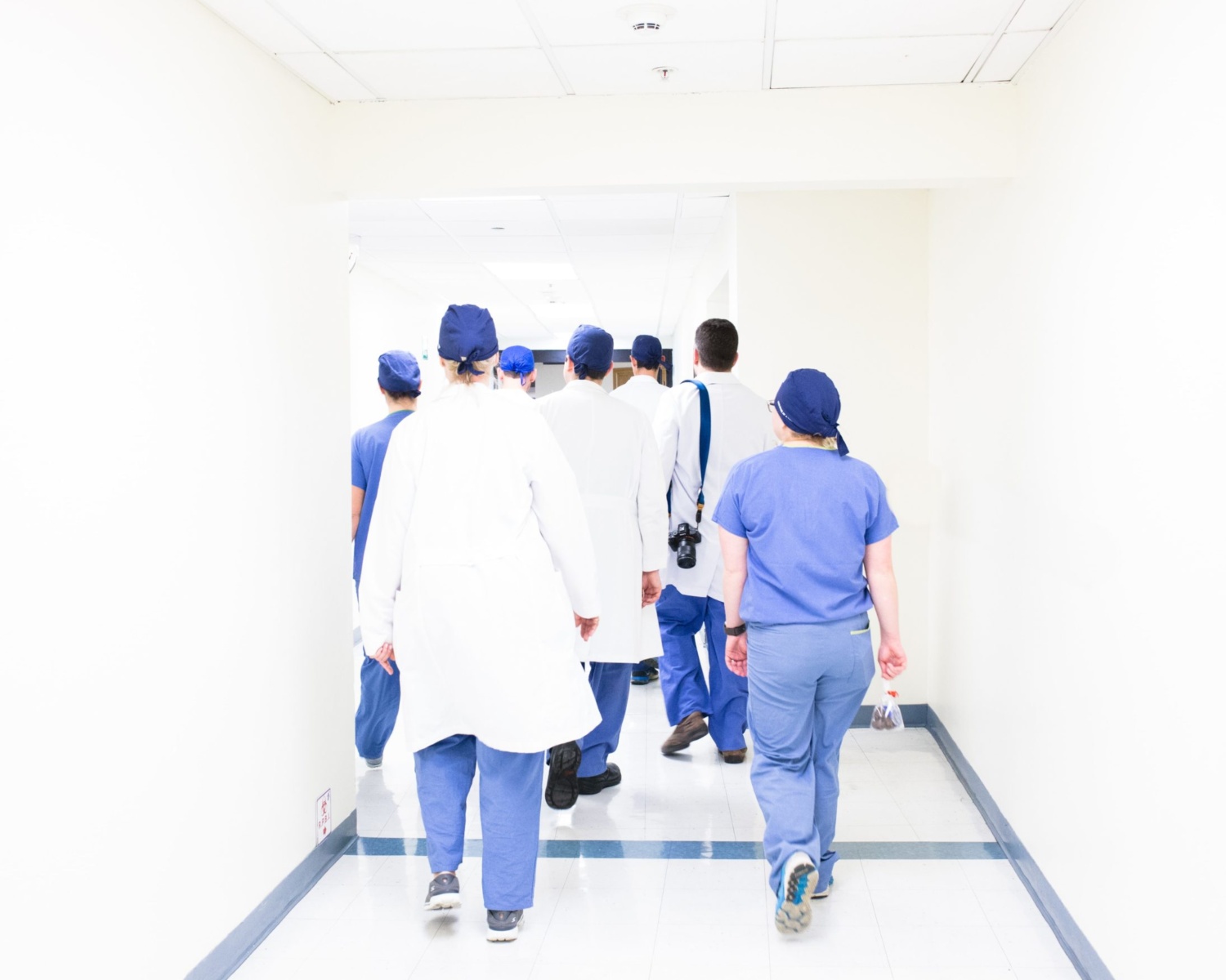 A group of doctors and nurses wearing blue scrubs and white coats walk away from the camera down a bright, white hospital hallway.