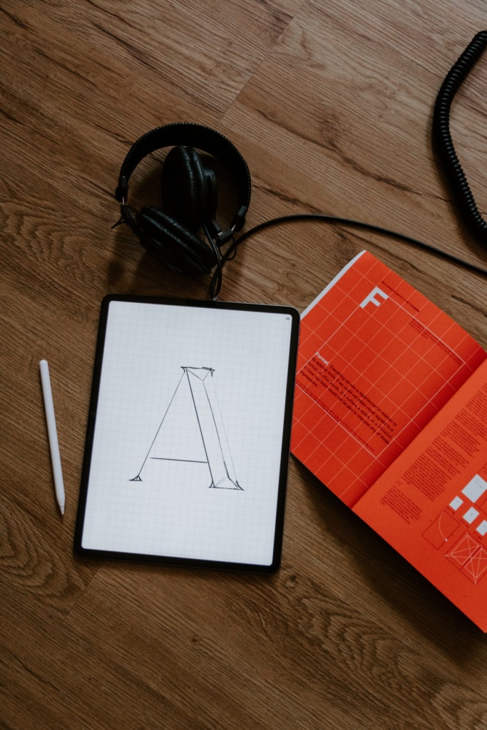 Typography sketch on tablet with design references Digital tablet showing a sketch of the letter “A” next to a stylus, headphones, and an open typography design book.